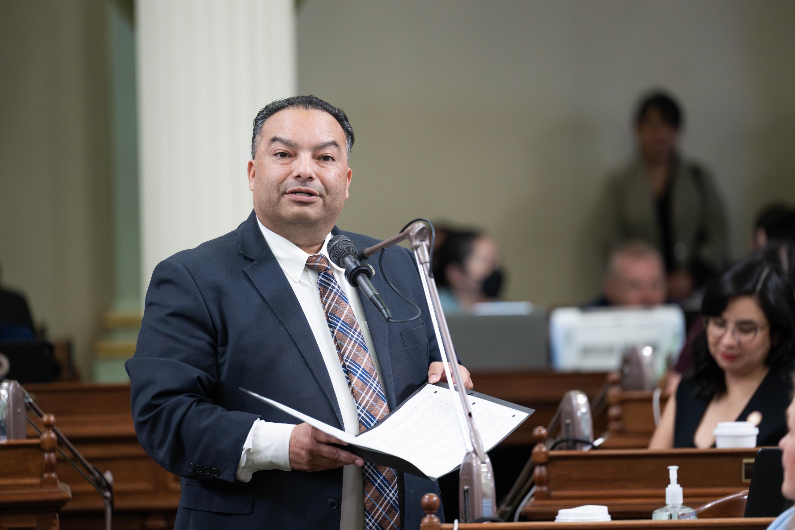 Asm. Solache standing at microphone on the Assembly Floor, speaking and holding folder