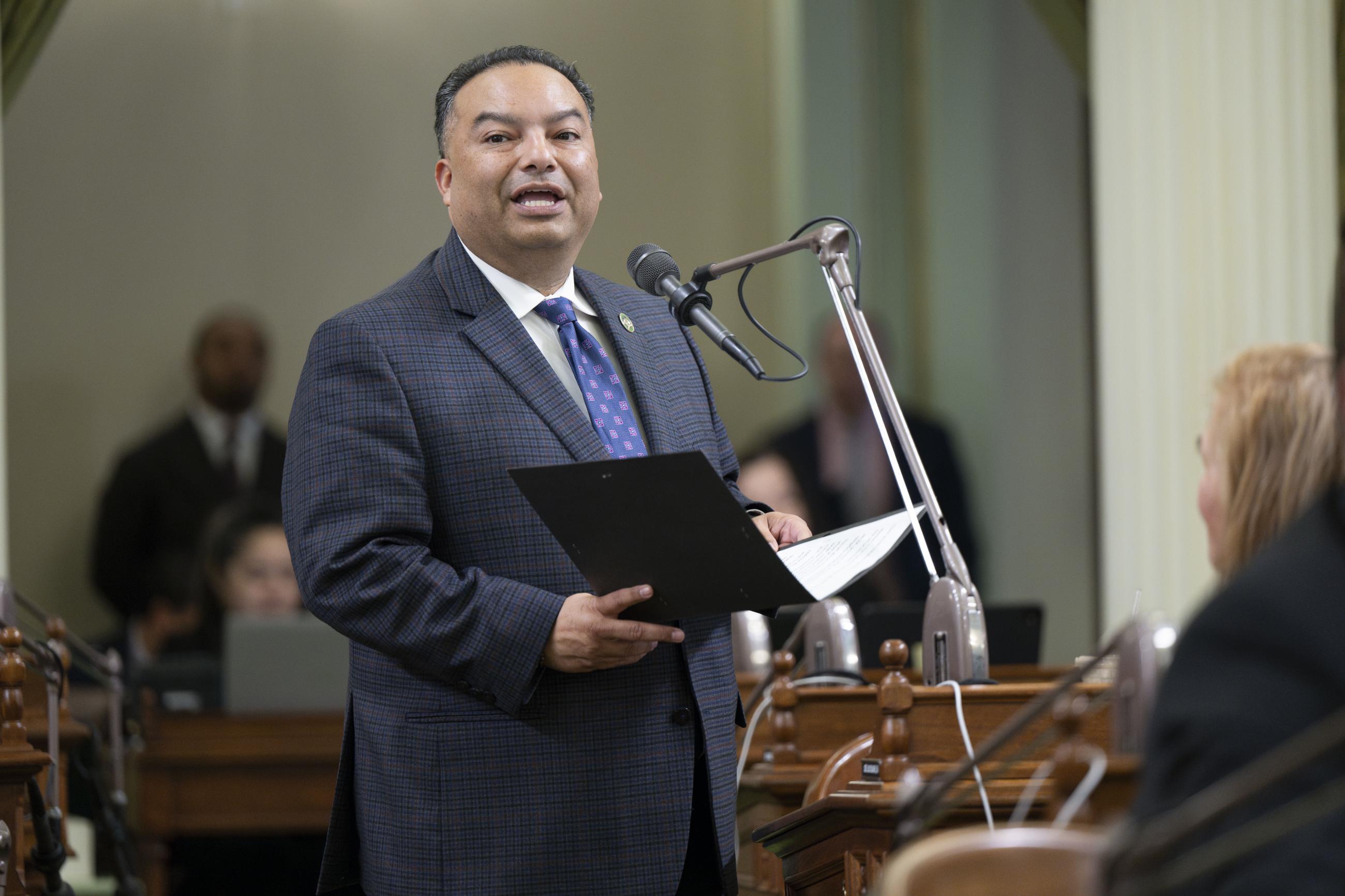 Asm. Solache standing at microphone, speaking and holding folder
