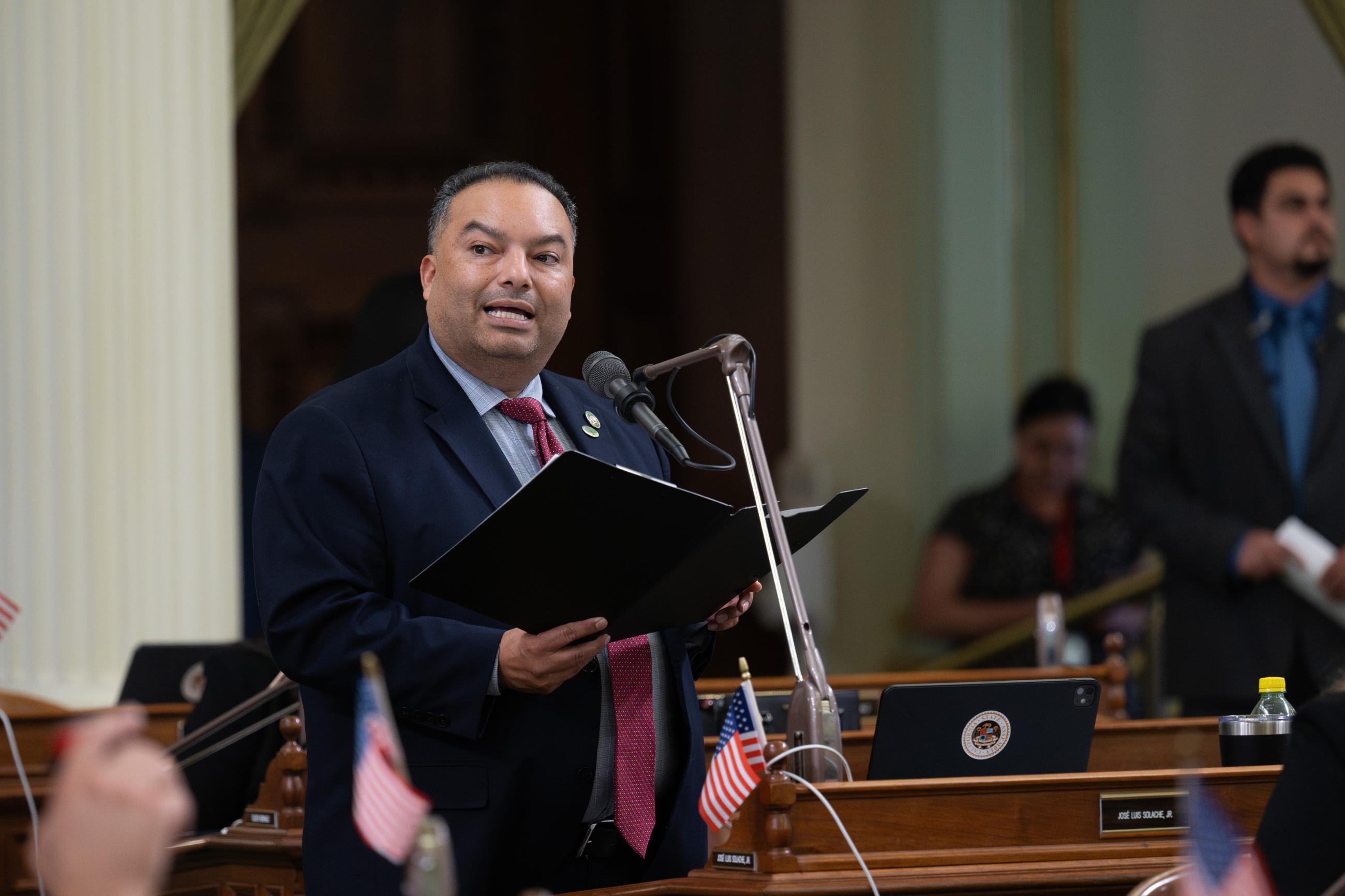 Asm. Solache standing at microphone, speaking and holding folder