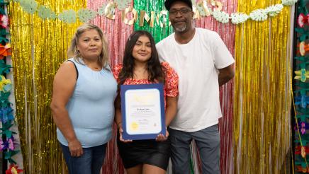 Student holding up certificate, with parents, in front of festive background