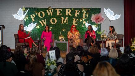 Guest speaking panel seated, with large "Powerful Women" sign in background