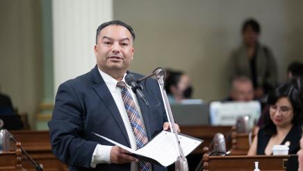 Asm. Solache standing at microphone on the Assembly Floor, speaking and holding folder