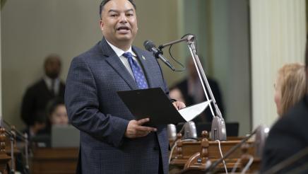 Asm. Solache standing at microphone, speaking and holding folder