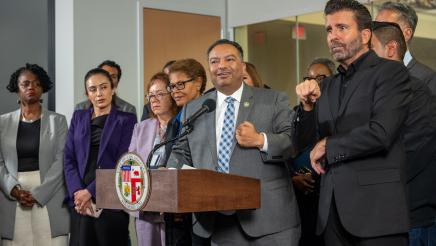 Asm. Solache at podium, speaking and gesturing with one hand, as Mayor Bass and others listen on