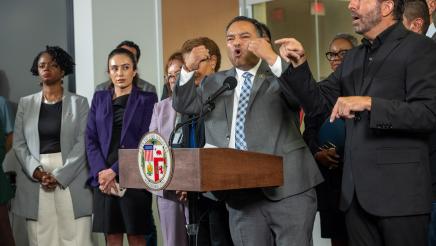 Asm. Solache at podium, speaking and gesturing with both hands, as Mayor Bass and others listen on