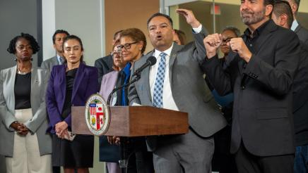 Asm. Solache at podium, speaking and gesturing with one hand, as Mayor Bass and others listen on
