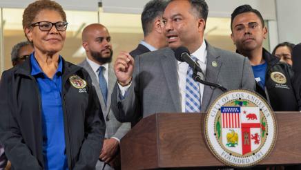 Asm. Solache at podium, speaking and holding up one hand, as Mayor Bass and others listen on