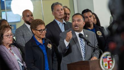 Asm. Solache at podium, speaking and holding up a fist, as Mayor Bass and others listen on