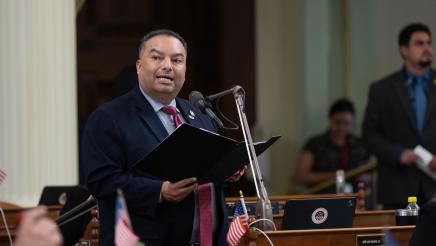 Asm. Solache standing at microphone, speaking and holding folder